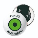 Imagen de un broche. En primer plano, puede verse un broche blanco circular de 7 cm de diametro, Donde arriba en forma curva dice en letrs negras mayúsculs "Tengo" y debajo del mismo modo "Baja Visión". En el centro hay otro circuló de circunsferencia negra relleno de verde, con un punto circular negro en el centro, simbolizando un iris